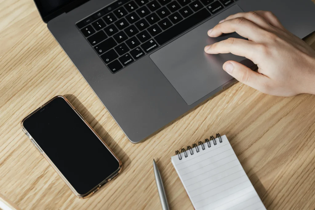 An overhead shot of a person's hand using the trackpad of a silver laptop on a light wooden desk, flanked by a black smartphone and a small spiral notepad with a silver pen.